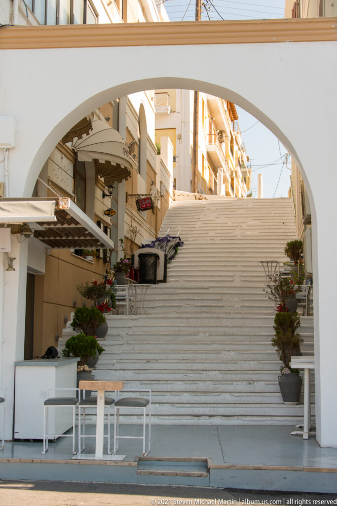 Wide white staircase climbing beneath an arch between buildings, with café tables, planters, balconies, and awnings.