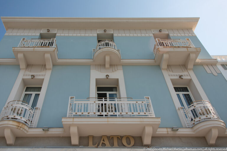 Upward view of pale blue LATO façade with white projecting balconies, curved corner railings, and the building name at street level.
