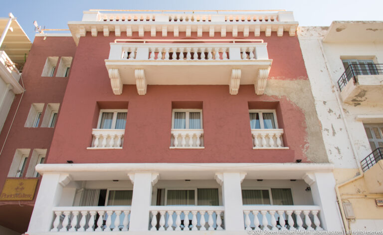 Red multi-story façade with white balustraded balconies, projecting cornices, columns, and repeated rectangular windows.
