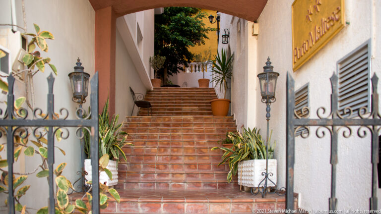 Terracotta-tiled steps rising through an open wrought-iron gate, flanked by lanterns and potted plants at a hotel entrance.