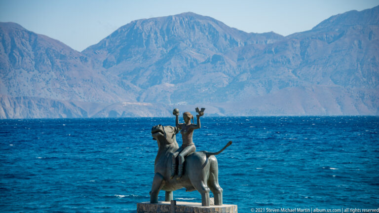 Seafront sculpture of a human figure riding a bull, silhouetted against blue water and rugged mountains across Mirabello Bay.