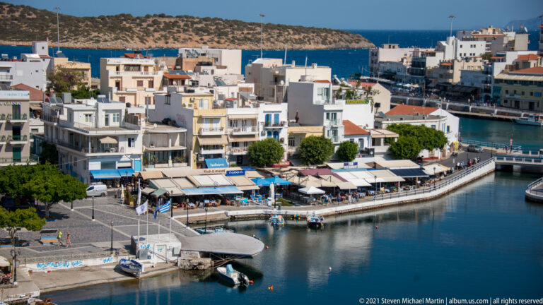 Curving Agios Nikolaos waterfront with restaurants, apartment buildings, promenade, moored boats, and blue sea beyond.