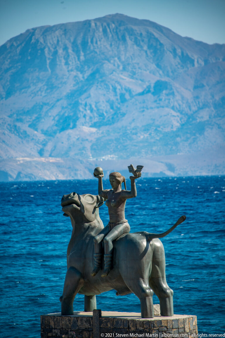 Side view of a bull-and-rider sculpture beside Mirabello Bay, with pale rugged mountains spanning the horizon.