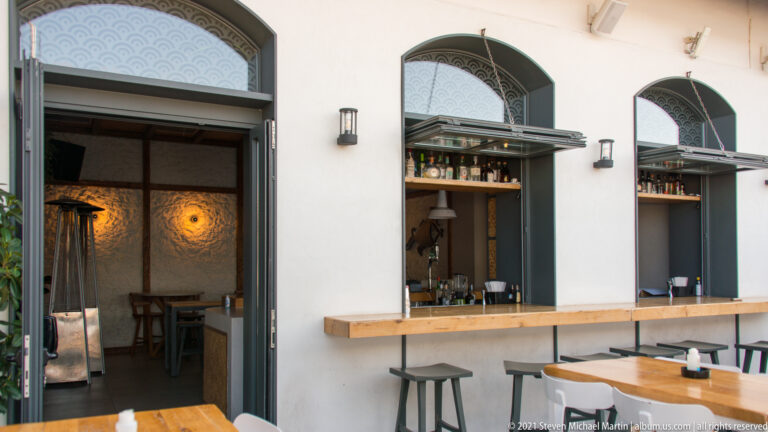 White café-bar façade with tall arched openings, open service windows, wooden counter, stools, and warm interior lighting.