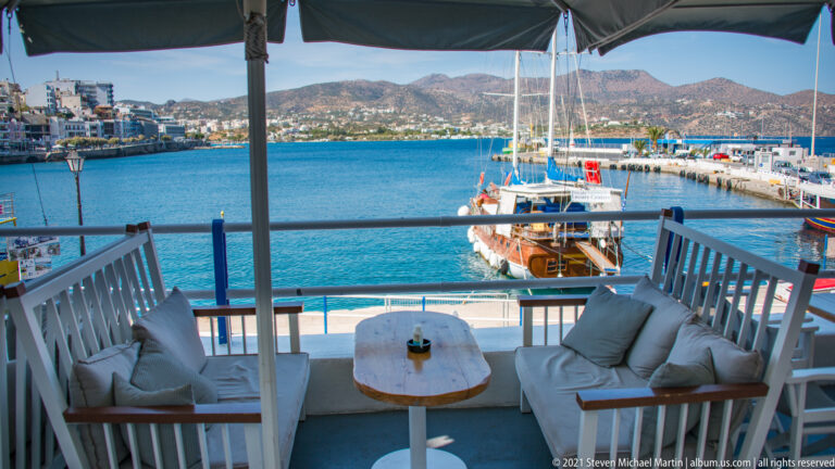 Cushioned lounge terrace under shade overlooking a moored sailing vessel, blue marina water, town, and dry hills.
