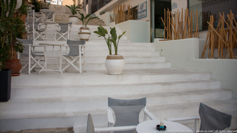 Whitewashed stepped café terrace with white chairs and tables, large potted plants, and reed screens.