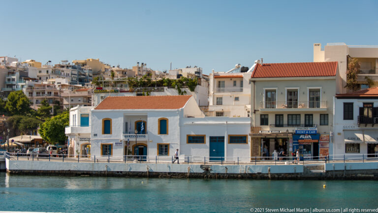 White and pastel waterfront buildings and shops along an Agios Nikolaos quay, with dense hillside apartments rising behind.