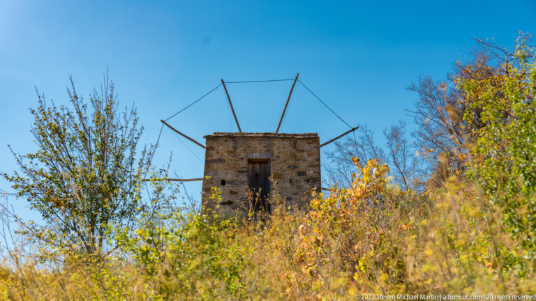 Weathered stone windmill tower among dry vegetation, with an exposed radial sail frame against a clear blue sky.