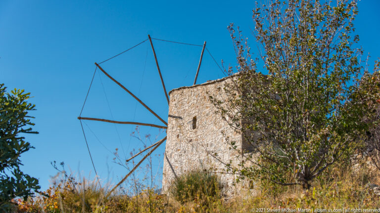 Oblique low-angle view of a stone windmill with bare radial sail supports, partly screened by a slender tree.
