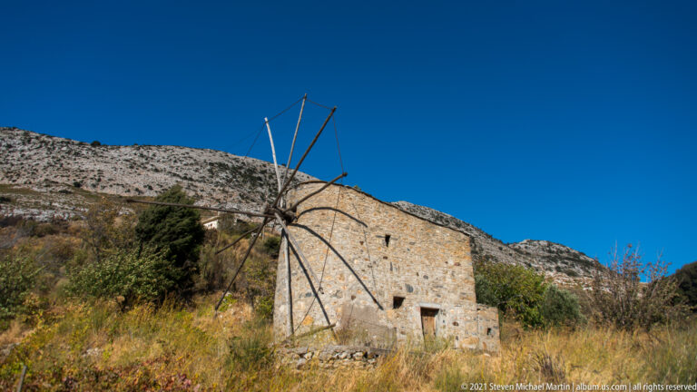 Stone windmill with bare radial sail frame on a dry hillside, backed by a steep rocky mountain slope.