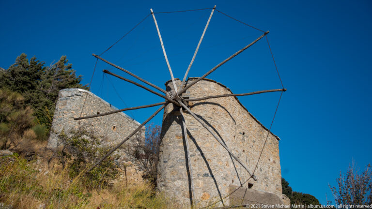 Low-angle close view of a stone windmill, with central hub and long bare sail spars extending across a deep blue sky.