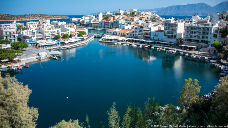 Wide view across Lake Voulismeni toward Agios Nikolaos waterfront buildings, with distant mountains under a clear blue sky.