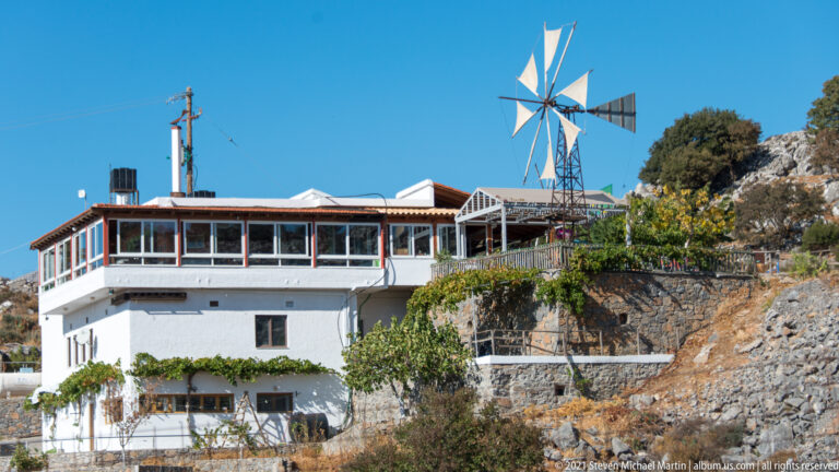 Metal multi-bladed wind pump on a lattice tower beside a white hillside building, rocky slope, and vines.