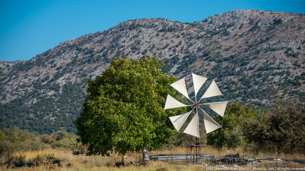Small multi-bladed wind pump in a dry field beside a leafy tree, with steep Lasithi mountains rising behind.
