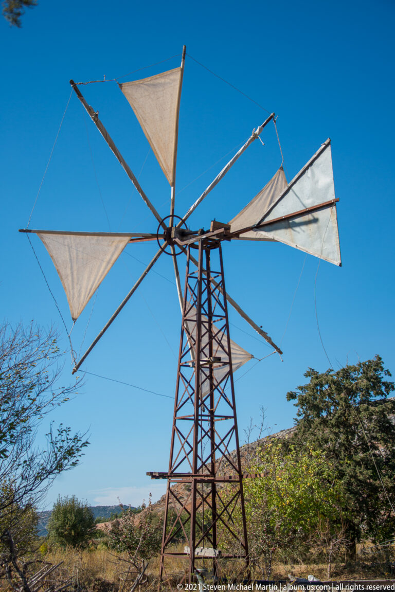 Metal-framed wind pump with triangular white fabric sails, long spars, cords, and a lattice support tower.