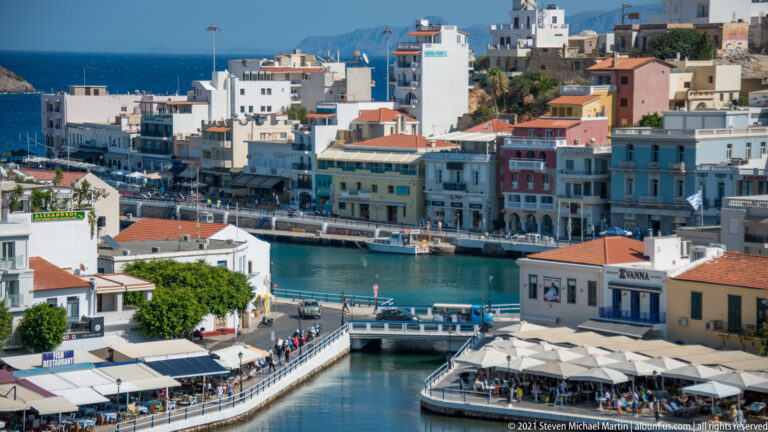Narrow harbor channel in Agios Nikolaos bordered by buildings, pedestrian quays, café terraces, and moored boats.
