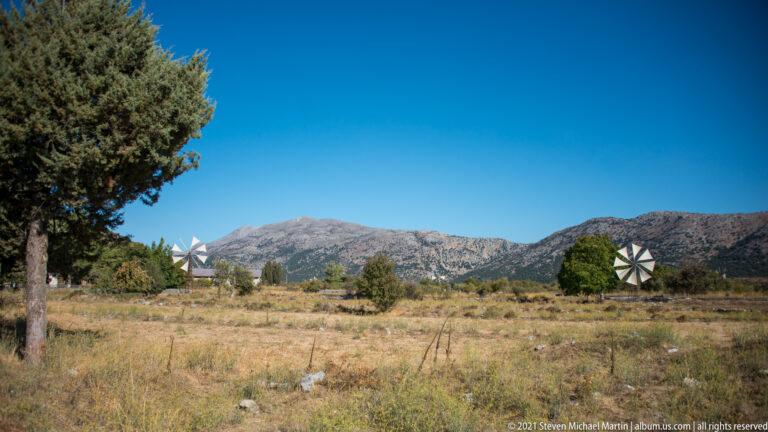Open Lasithi Plateau fields with several fabric-sailed wind pumps, scattered trees, and rugged mountains under a clear sky.