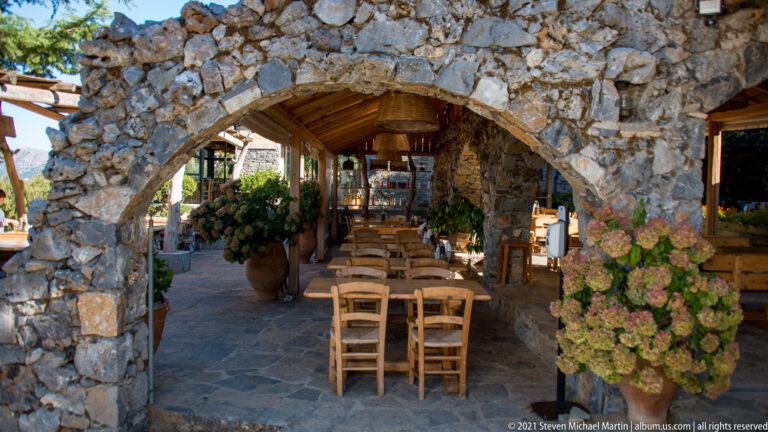 Rustic taverna dining area seen through a broad stone arch, with wooden tables, chairs, hydrangeas, and timber roof.