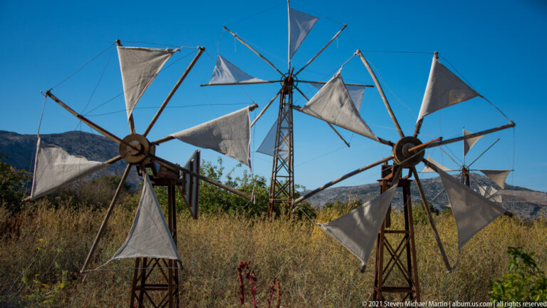 Three large fabric-sailed wind pumps among dry grasses, with additional wind pumps visible farther across the Lasithi Plateau.