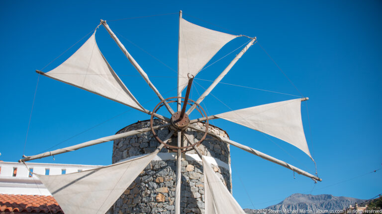 Stone tower windmill with triangular white fabric sails and long radial spars against a vivid blue sky.