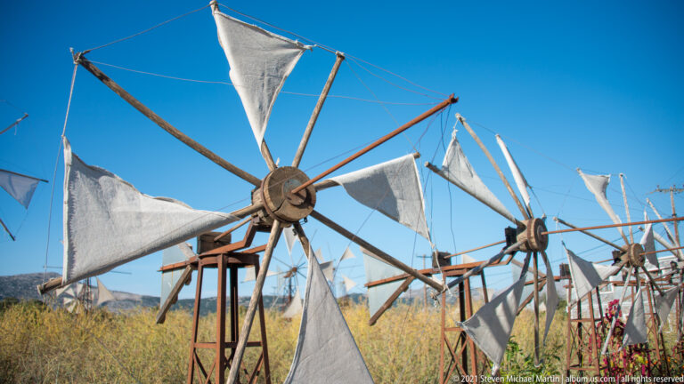 Row of fabric-sailed wind pumps extending across dry Lasithi fields, with one large rotor dominating the foreground.