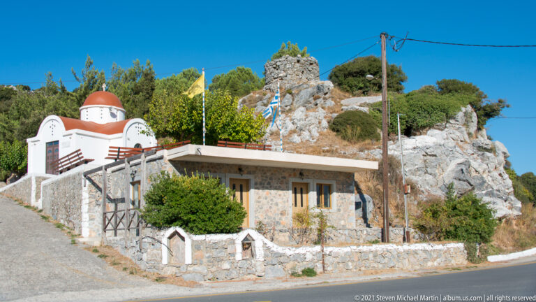 Small white Orthodox chapel with red dome, Greek flags, and stone annex beside a rocky roadside outcrop.