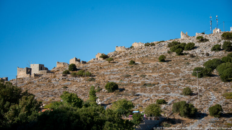 Rocky hillside dotted with ruined stone windmills and low masonry structures, with communication masts on the summit.