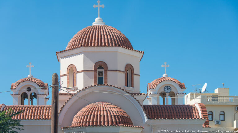 Terracotta domes, arched openings, crosses, and pale stucco walls of an Orthodox church against a bright blue sky.