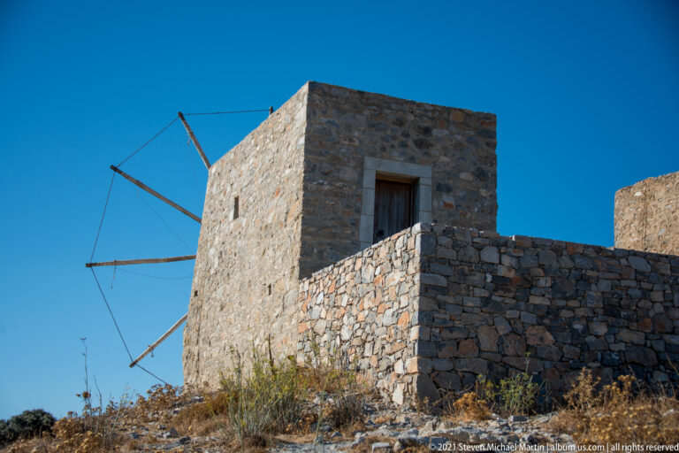 Large square stone windmill with tall doorway and bare radial sail spars on a dry rocky Lasithi hillside.