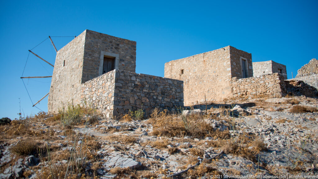 Group of weathered stone windmills on a rocky Lasithi slope, one retaining part of a radial sail framework.