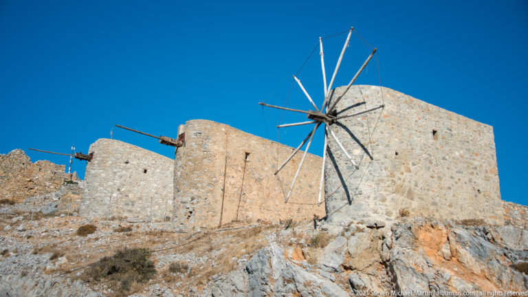 Three large stone windmills on a rocky slope, one with a prominent radial sail framework and others with projecting axles.