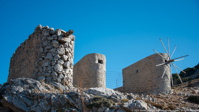 Ruined stone wall in front of two surviving windmill towers, one retaining a skeletal radial sail frame.