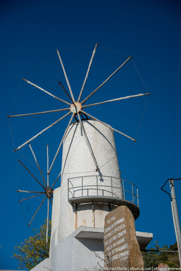 Whitewashed cylindrical windmill with bare radial sail spars and projecting balcony, with a second mill partly visible behind.
