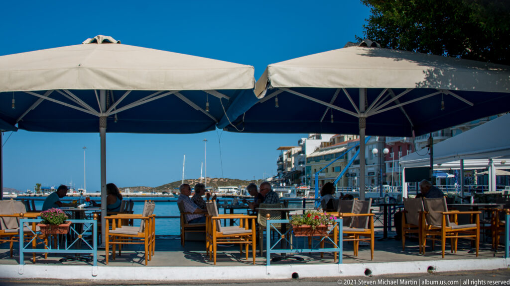People seated beneath large umbrellas at a waterfront café, with Agios Nikolaos harbor and boats visible beyond the railing.