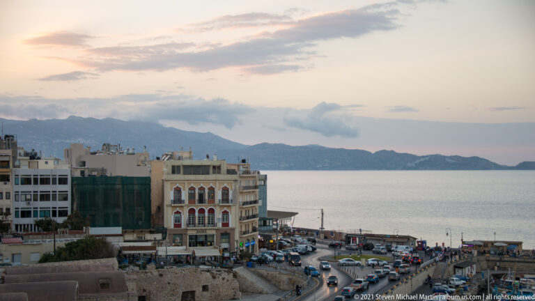 Elevated dusk view over Heraklion waterfront buildings and traffic toward the calm Cretan Sea and distant mountainous coast.