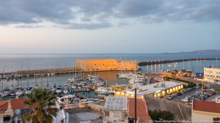 Illuminated Koules Fortress at Heraklion's harbor entrance, with marina masts, breakwater lights, and open sea beyond.