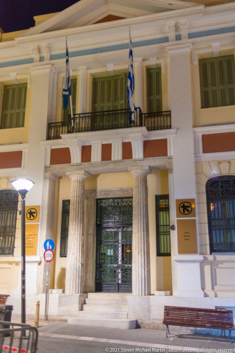 Night view of a pale civic building with columns, green shutters, balconies, and Greek and European Union flags above the entrance.