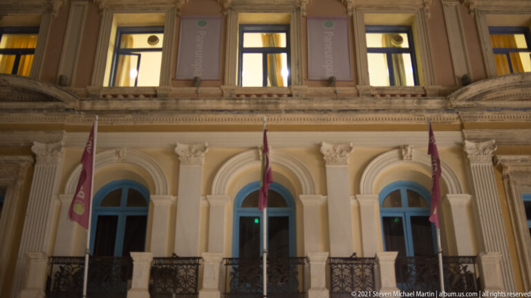 Night detail of the Heraklion Loggia with illuminated arches, columns, balustrade, flags, and ornate carved stonework.