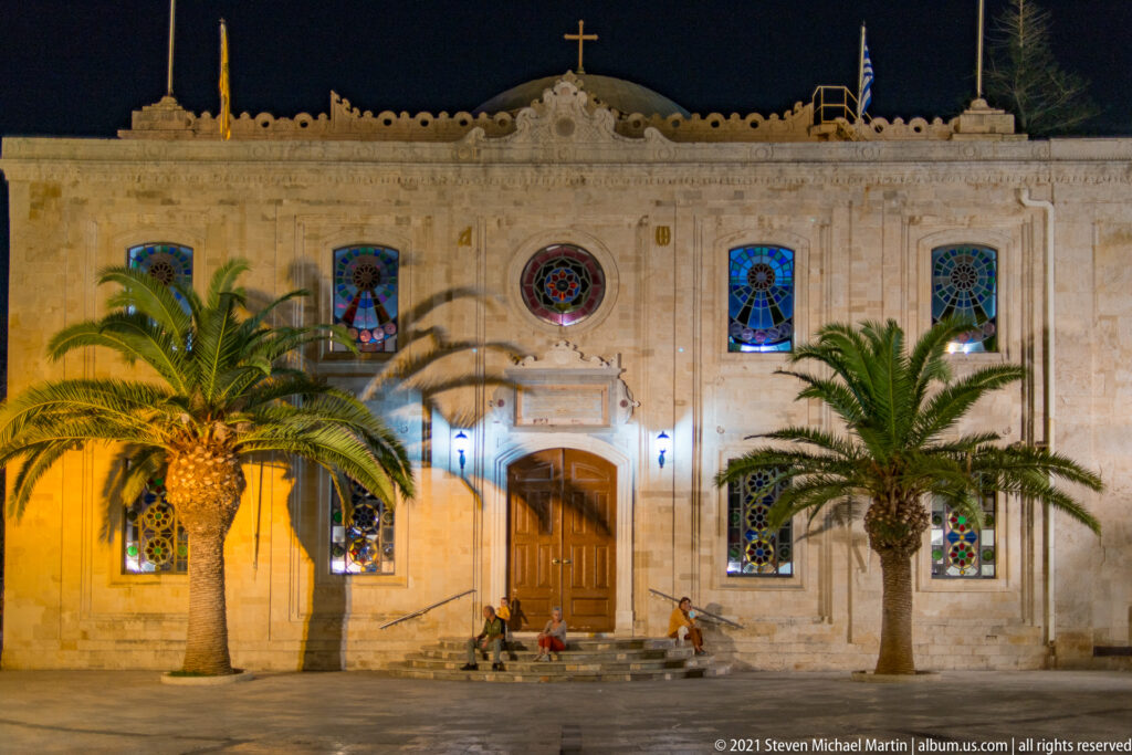 Illuminated Agios Titos Church with central wooden door, stained-glass windows, roof cross, and palm trees in the square.