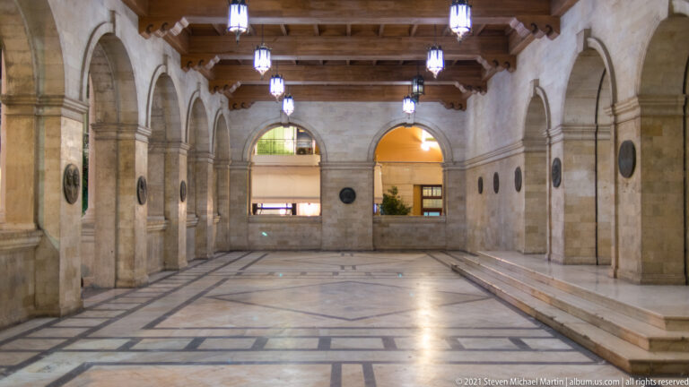Long interior hall of the Heraklion Loggia with stone arches, wall medallions, timber ceiling, hanging lights, and patterned marble floor.