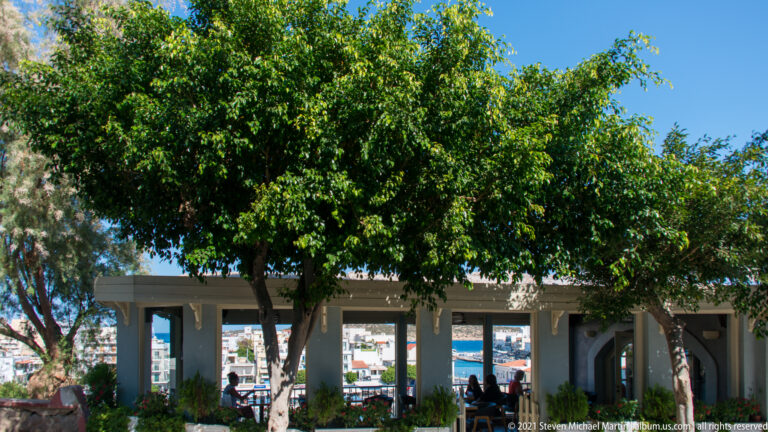 Leafy public terrace with trees, benches, and a low colonnade overlooking Lake Voulismeni and Agios Nikolaos.