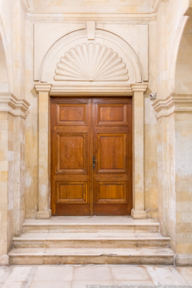 Double wooden doors reached by marble steps beneath a shell-headed stone arch inside the Heraklion Loggia.