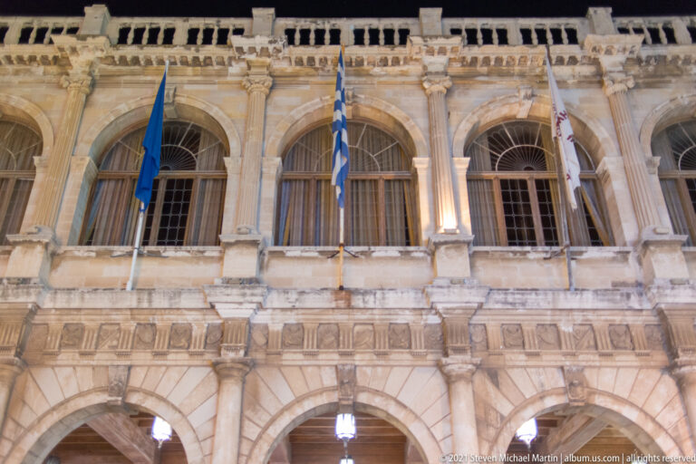 Night close-up of the Heraklion Loggia upper façade with large arches, carved balustrade, and Greek, EU, and civic flags.