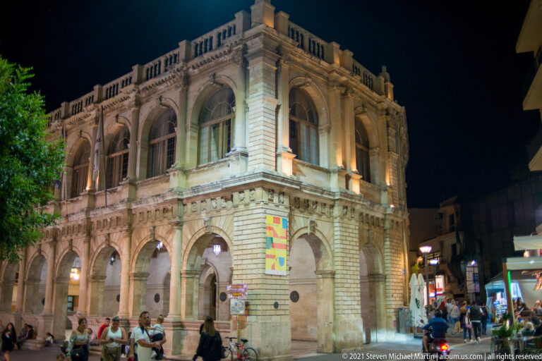 Illuminated Heraklion Loggia at night, with ground-floor arcades, tall upper arches, roof balustrade, and pedestrians around the corner.