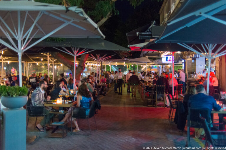 Busy Heraklion outdoor café terrace at night with large umbrellas, crowded tables, trees, and colorful restaurant lighting.