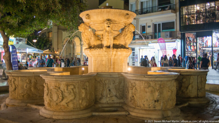 Illuminated Morosini Lions Fountain with carved stone basins, four lion heads, flowing water, and pedestrians in Heraklion's Lions Square.