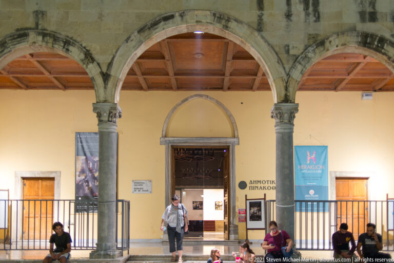Arched entrance to Heraklion Municipal Art Gallery with Greek gallery signage, exhibition banners, visitors, and stone columns.