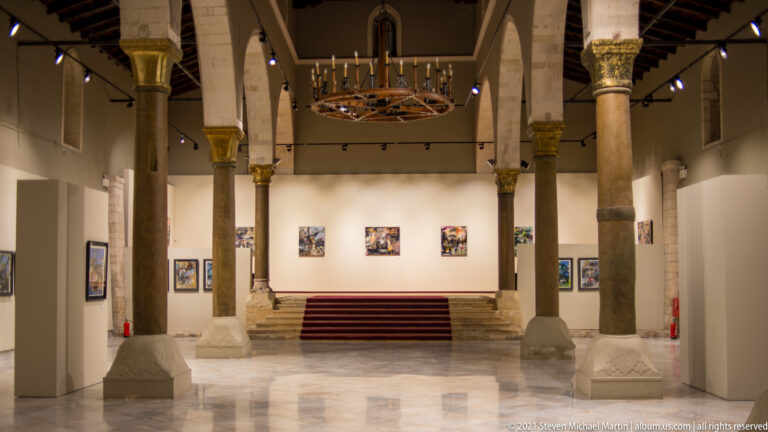 Interior of Heraklion Municipal Art Gallery with stone columns and pointed arches, timber roof, circular chandelier, and artworks on the walls.