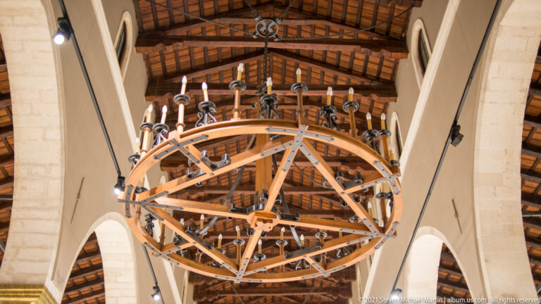 Large circular ring chandelier with many candle-like lights suspended beneath timber roof beams between stone arches.