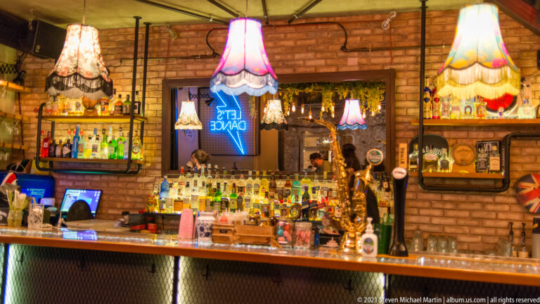 Eclectic cocktail bar with colorful fringed lamps, rows of bottles, brick walls, mirrors, and a blue LET'S DANCE neon sign.